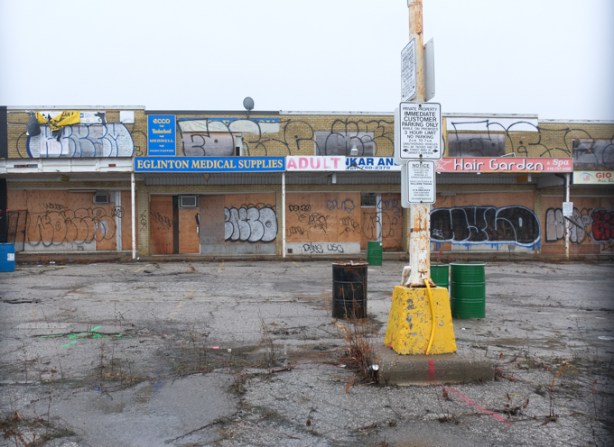 boarded up and tagged over, old lowrise strip mall at Kennedy and Eglinton Adult store, Eglinton Medical supply, and Hair Garden, cracked pavement in front,