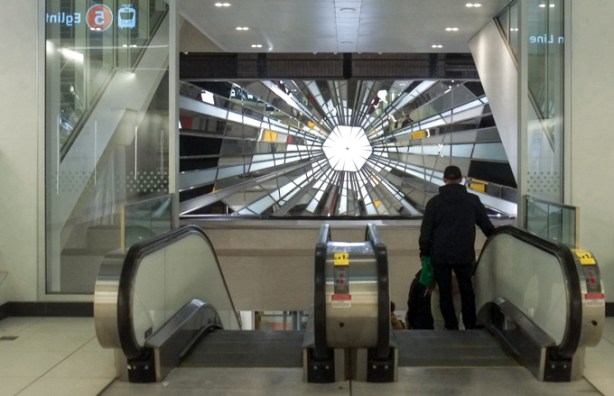 man going down escalator at Eglinton station, wall in front of him has artwork made of pieces of glass and mirror that look llke something has smashed through the center of a mirror or window. 