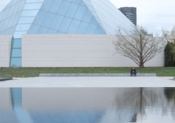 person sitting on bench by reflecting pool at aga khan museum, on phone, lots of reflections in the water and on glass of islamic center