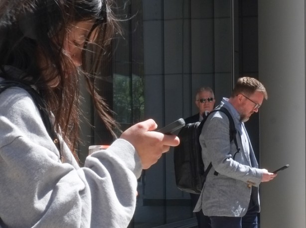 three people, two on phones and a third watching. One is a woman with long hair, the others are men