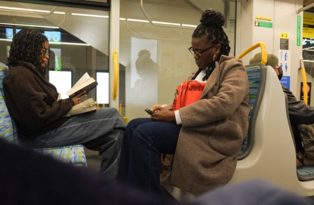 women riding on subway, one is reading a book and the other is on her phone