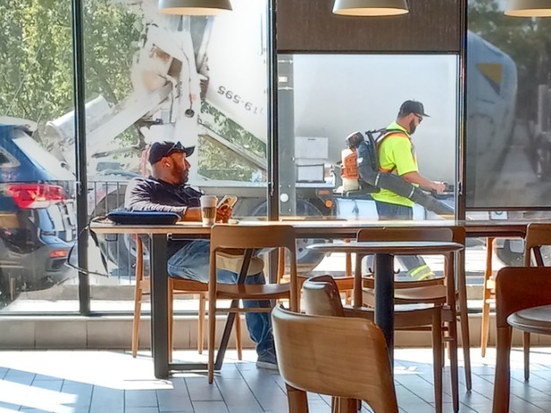 man at table with phone, inside at Starbucks, beside large window, another man with a leaf blower is working outside