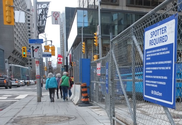 gray haired senior women walking west on bloor towards yonge intersection, with construction fence beside them, north side of bloor