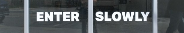the words enter slowly on the glass portion of a garage door, with reflections in that glass of people walking past