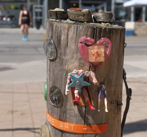 old wood stump, upright on sidewalk, with plastic figure nailed to it as artwork
