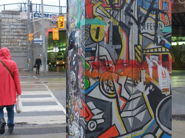 person in red rain coat crossing queen street west at dufferin, painted sidewalk box in abstract design in the foreground, railway underpass with traffic in the background