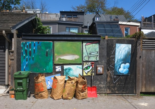 on a fence in an alley, part of the fence is covered with 5 or 6 paintings, bags of lawn garbage lined up in front of the fence, back of houses behind