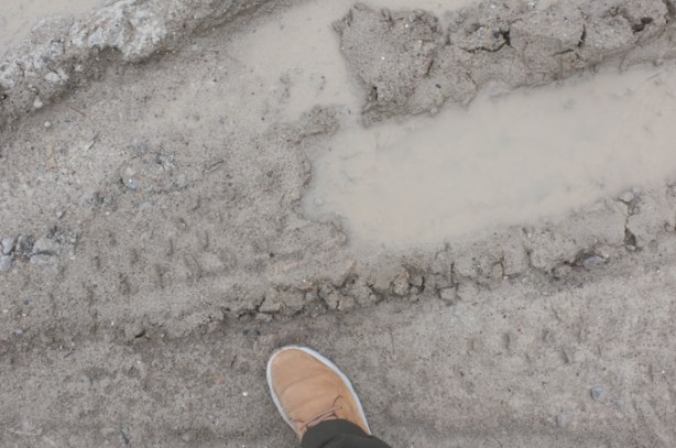 looking down, foot (with shoe) stepping beside muddy puddle and tire tracks in the mud