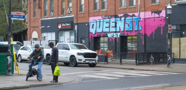 two men on a corner, across from building with mural on exterior wall, says queen street west in blue letters on pink background, 