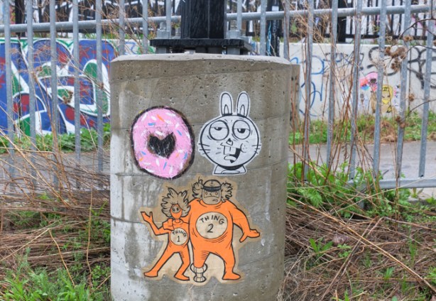 three paste up graffiti pieces on a concrete support to a pole. One is T bonez and trp 613 sailor dude dressed as thing 1 and thing 2 from Dr Suess Cat in the hat. second paste up is pink donut with a heart shaped hole and last is a white rabbit face