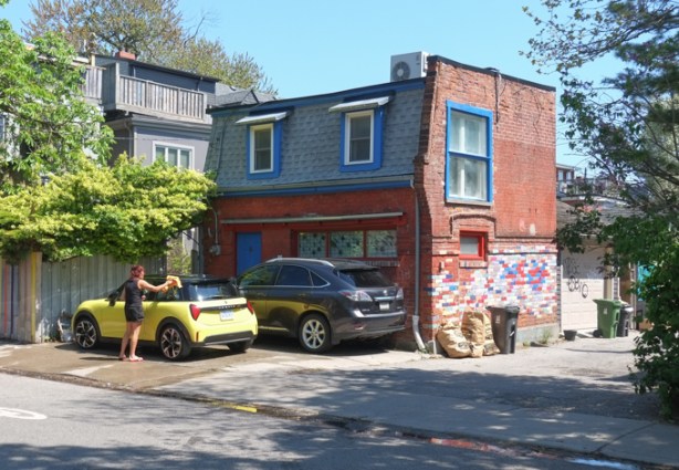 a woman is outside, washing a yellow car, in front of a house with a mansard roof,