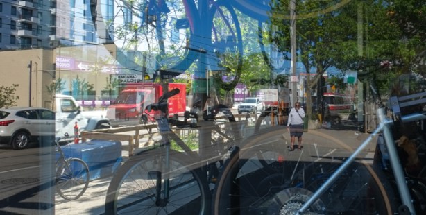 looking through a bicycle store window with man walking on sidewalk, a bus, and other street scene, bikes in the store and hanging from the ceiling