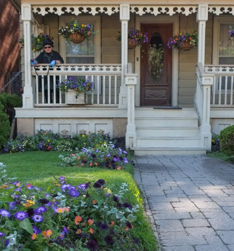 woman standing on front porch with a hose, watering garden of flowers