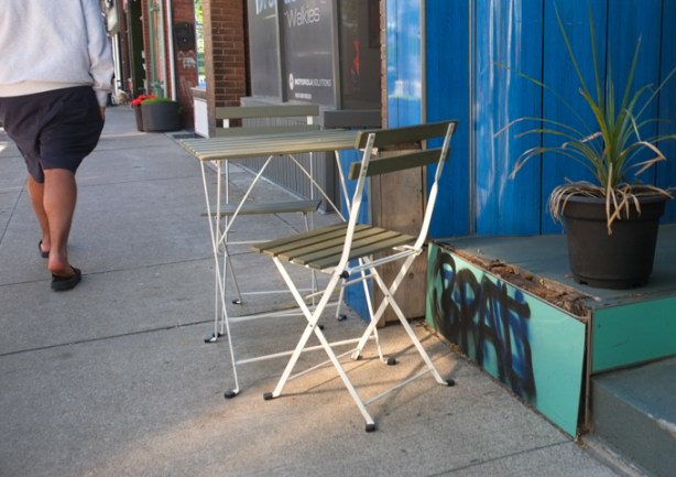 a man in shorts and sandals walks past a small table and two olive green chairs on the sidewalk