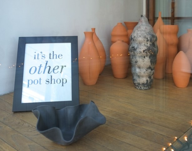 in the window of a pottery store, some terra cotta pots, also a sign that says it's the other pot shop