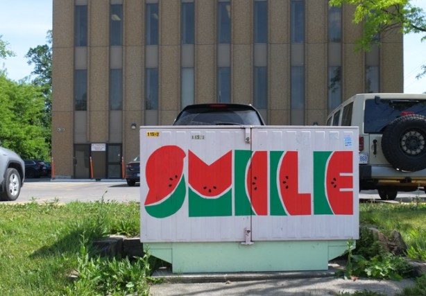 metal bell box on sidewalk painted with the word smile such that letters look like slices of watermelon