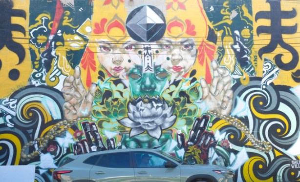 a car is parked in front of part of a mural by flips and rowell soller, 2 white faces, with a green face in the middle, yellow curvy curls