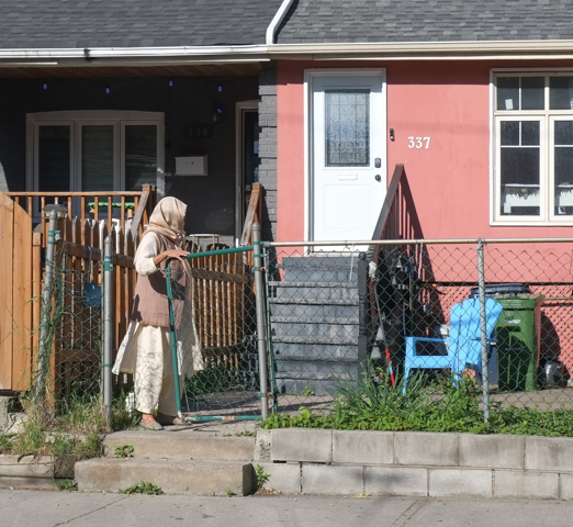 a woman wearing a head scarf is opening front gate in front of a small orange house with a white door