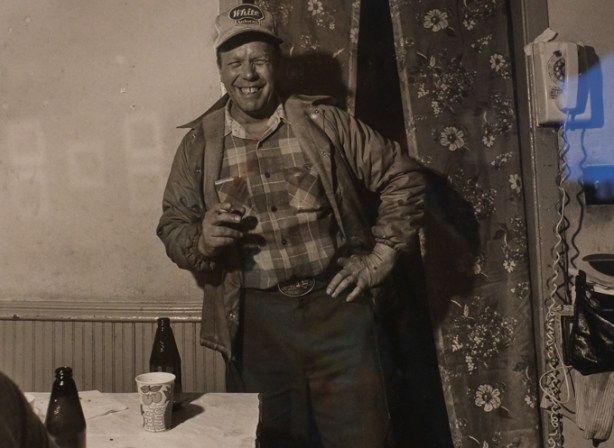 black and white photo by Larry Fink from his Social Graces series from the 1970s, man in plaid shirt, jacket, and baseball cap, is holding a drink. Eyes closed, but smiling,