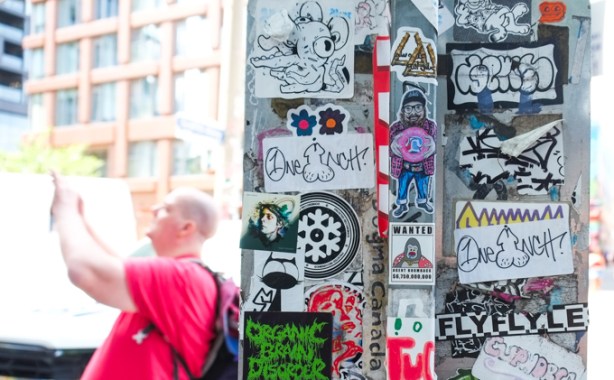 man in red shirt is taking a picture, behind him is a sign that is covered with graffiti stickers