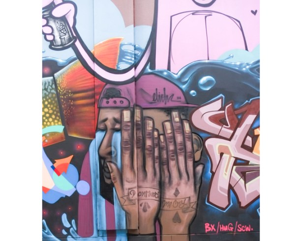 street art painting by elicser elliott, part of a larger collaborative mural, man with burgundy baseball cap, with hands over his eyes. back of hands have tattoo words, Don't watch me cry, written over the two hands