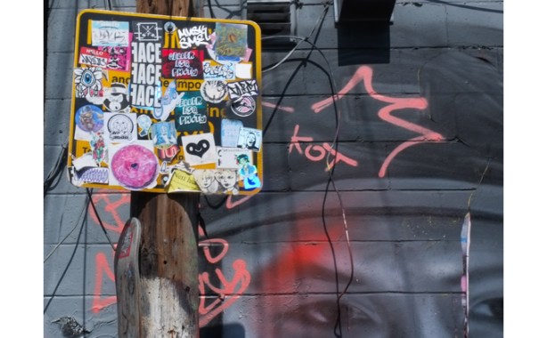 graffiti stickers covering a sign, a mural of a face is behind it