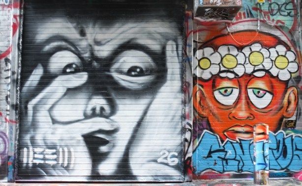 two graffiti paintings, on left is black and white face with fingers in front, on right is an orange face with a garland of daisies on its head