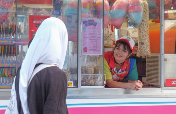 a young woman looks out the window of a food concession stand that sells cotton candy, candy apples, large lollipops, and popcorn, at the ex, she is smiling at another woman who is wearing a white head scarf