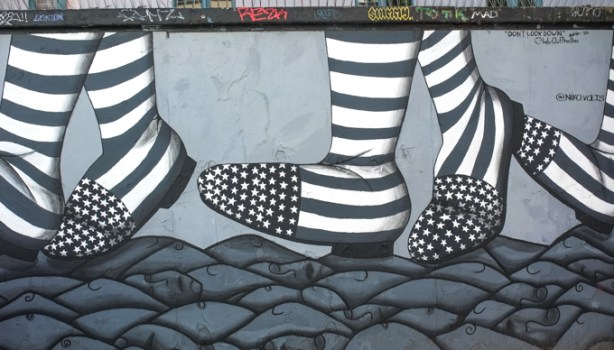 part of a mural in shades of grey, black and white, of feet wearing boots that look like the American flag, walking over the heads and faces of other people lying on the ground