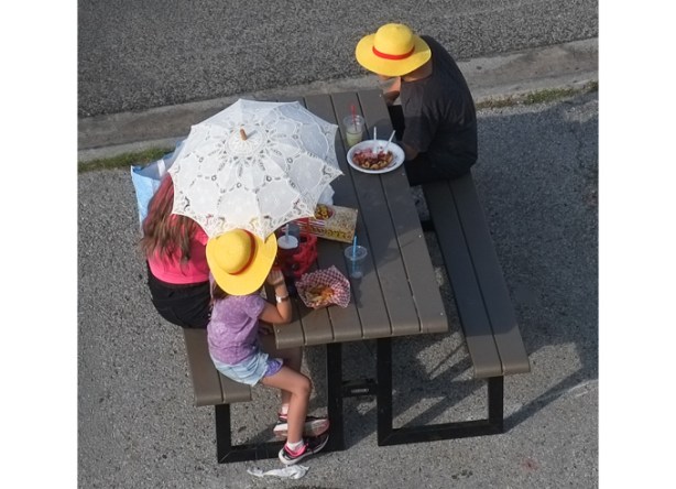 a hite parasol being held by a family eating at a picnic table at the ex, two of them are wearing yellow hats with red hatbands