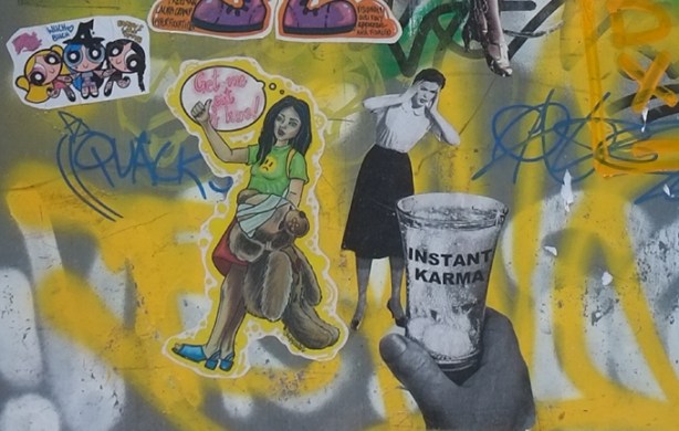 two graffiti paste ups, one is a young woman saying get me out of here, the other is a large hand holding a glass that says instant karma