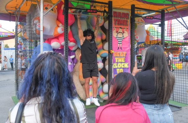 backs of heads of people watching a man trying to hold himself up, hanging onto a bar for more than two minutes, Canadian National Exhhibtion, midway