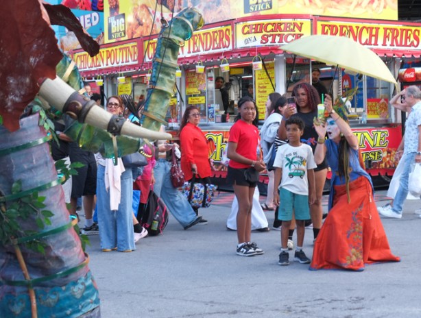 woman with umbrella and long red skirt, crouching down and talking to a boy as she holds a microphone in her hand, Canadian National Exhibition, midway