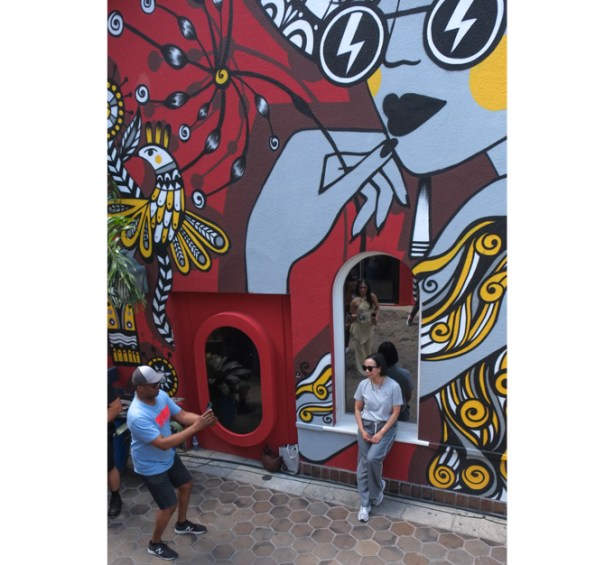 man taking picture of woman who is leaning standing beside wall with large mural by Ola Volo