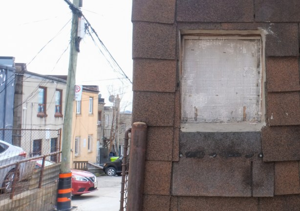 small window on a brown shingle covered shed, in an alley, back of stores on other side of the lane in the background